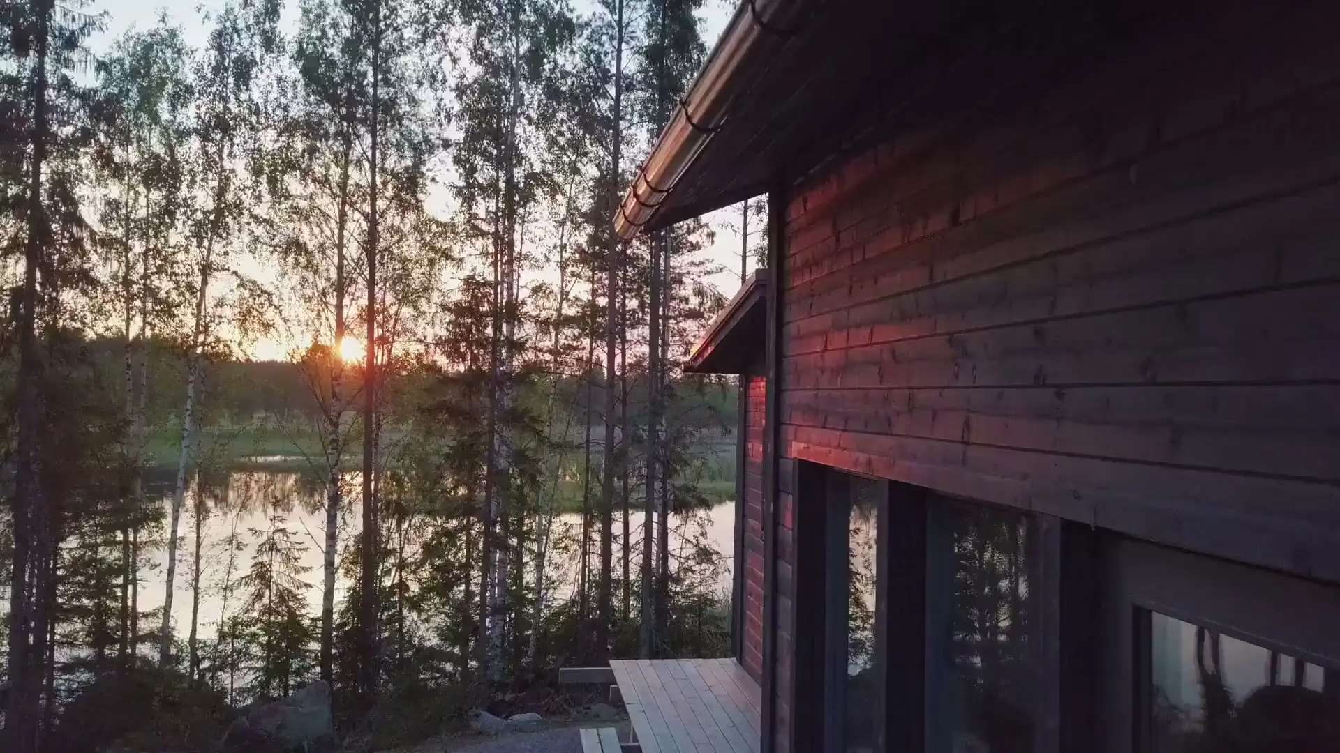 A dark timber lake house at sunset, seen through tall birch and pine trees with the sun low over calm water